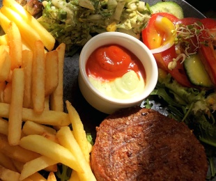 Zdjęcie przedstawia talerz, na nim Beyond Burger podany z frytkami, surówką, warzywami i sosami